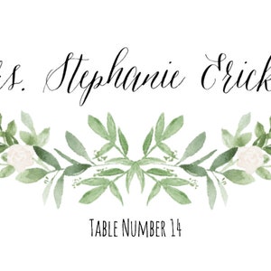 May include: A white card with black calligraphy that reads "Mrs. Stephanie Erickson" and "Table Number 14". The card is decorated with a green and white floral design.