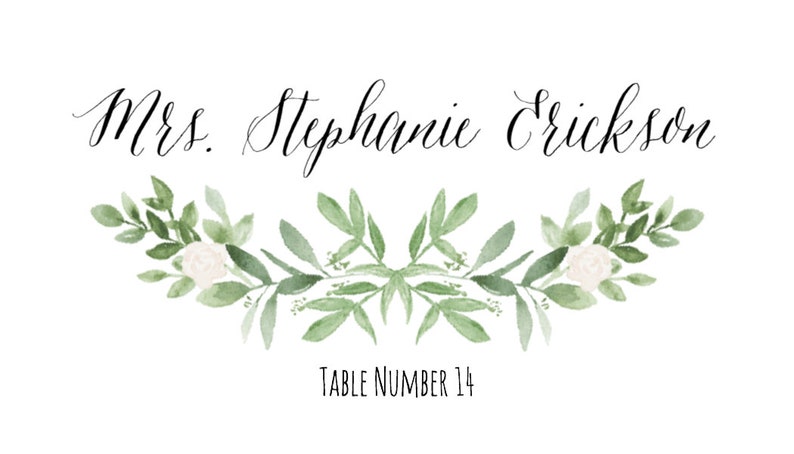 May include: A white card with black calligraphy that reads "Mrs. Stephanie Erickson" and "Table Number 14". The card is decorated with a green and white floral design.