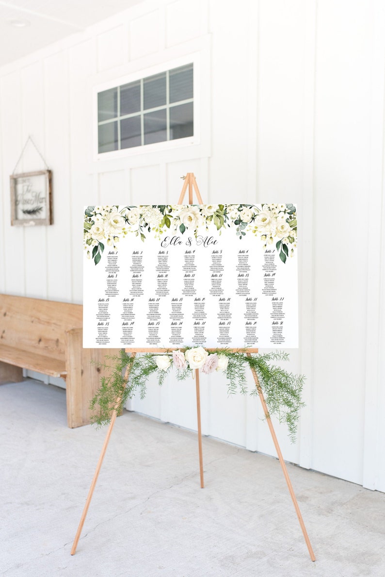May include: White wedding seating chart with green foliage and white flowers. The chart is on an easel with a floral garland. The chart lists table numbers and guest names. The chart reads "Ella & Alex".