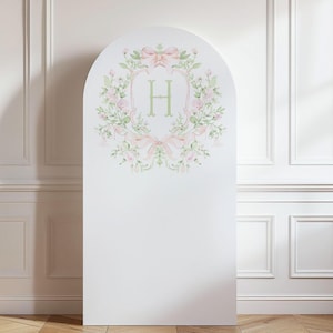 May include: White arched backdrop featuring a floral wreath design with pink bows and green foliage. The wreath surrounds a light green letter "H". The backdrop is set against a white wall with wooden flooring.