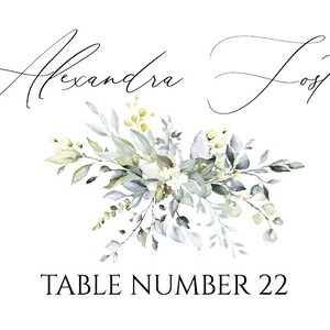 May include: A white card with a floral design in shades of green and white. The text "Alexandra Foster" is written in cursive at the top of the card. The text "TABLE NUMBER 22" is written in black at the bottom of the card.