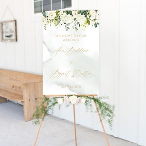 May include: A white wedding welcome sign with green foliage and white flowers. The sign reads "Welcome to our wedding, Ava Baldwin & Bryant Dalton, May 22, 2021".