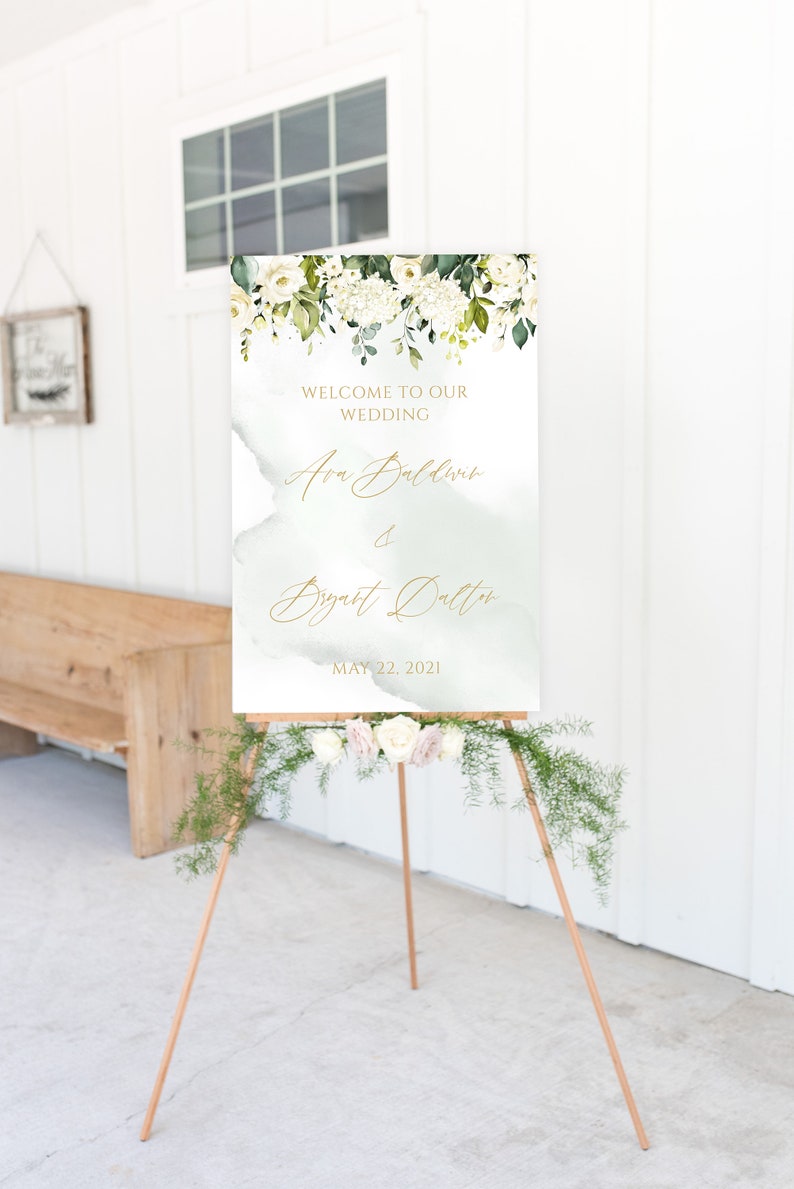 May include: A white wedding welcome sign with green foliage and white flowers. The sign reads "Welcome to our wedding, Ava Baldwin & Bryant Dalton, May 22, 2021".
