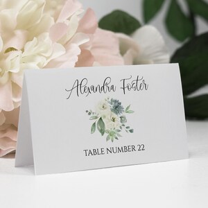 May include: White place card with black text that reads "Alexandra Foster" and "Table Number 22". A watercolor illustration of a bouquet of white and blue flowers with green leaves is centered above the text.
