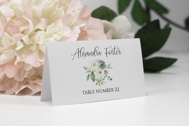 May include: White place card with black text that reads "Alexandra Foster" and "Table Number 22". A watercolor illustration of a bouquet of white and blue flowers with green leaves is centered above the text.