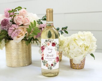Puede incluir: Una botella de vino de vidrio transparente con tapa dorada y una etiqueta blanca con una corona floral de hojas rosas, rojas y verdes. El texto de la etiqueta dice Avery and Bennett, To Have and to Hold, 21.3.2019 en fuente negra.