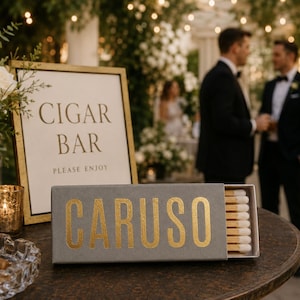 May include: A rectangular grey matchbox with the word CARUSO printed in gold lettering on the front. The matchbox is partially open, revealing a row of wooden matches with white tips. The box is positioned on a dark surface next to a cigar bar sign.