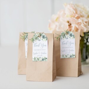 May include: Three brown paper bags with white thank you tags. The tags have a floral design and the text "Thank You for celebrating with us Sophia & Graham May 21, 2023".