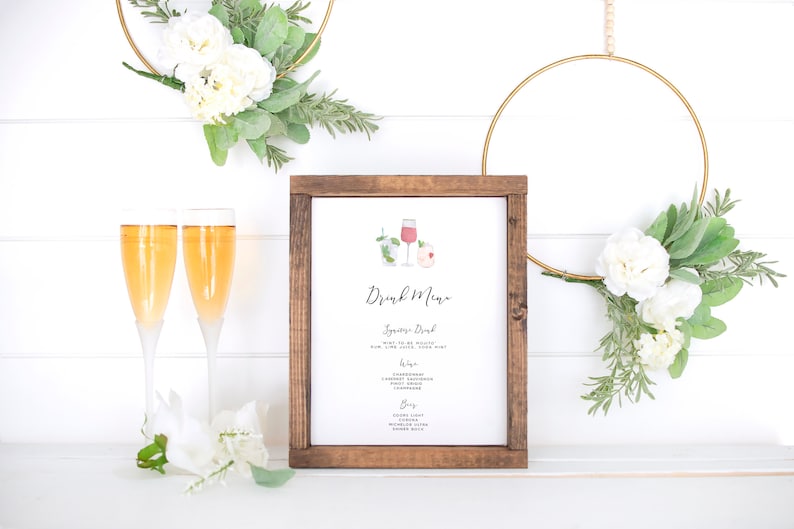May include: A wooden framed sign with a white background and the text "Drink Menu" in black calligraphy. The sign features two watercolor illustrations of cocktails, one with a pink drink and the other with a green drink. The sign is sitting on a white surface with two champagne flutes and a floral arrangement.