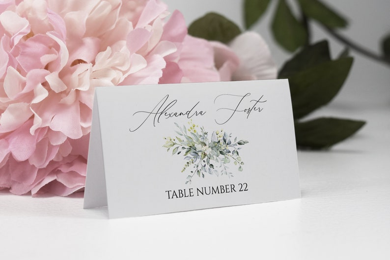 May include: White place card with a green floral design and the text "Alexandra Foster" and "Table Number 22".