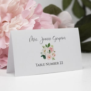 May include: White place card with a floral design and the text "Mrs. Joanne Simpson" and "Table Number 22".