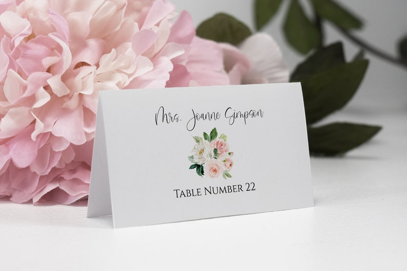 May include: White place card with a floral design and the text "Mrs. Joanne Simpson" and "Table Number 22".