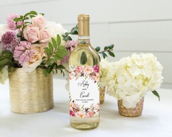 Puede incluir: Una botella de vino de cristal transparente con una etiqueta rectangular personalizada con un borde floral en tonos rosa y burdeos. La etiqueta muestra los nombres Ashley y Everett en letra negra, con el texto FROM THIS DAY FORWARD 11.9.2018.
