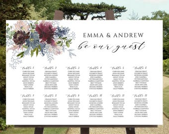 May include: A white wedding seating chart sign featuring a floral arrangement of burgundy, blue, and blush flowers in the top left corner. The text reads EMMA & ANDREW, be our guest, and lists guest names under twelve table headings.