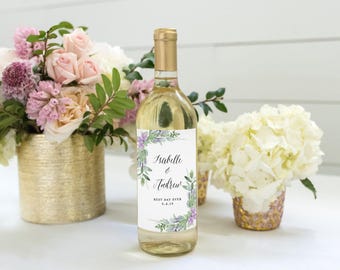 Può includere: Una bottiglia di vino in vetro trasparente con un'etichetta rettangolare bianca caratterizzata da un bordo floreale ad acquerello nei toni del viola e del verde. L'etichetta riporta il testo Isabelle & Andrew, BEST DAY EVER e 5.4.19.