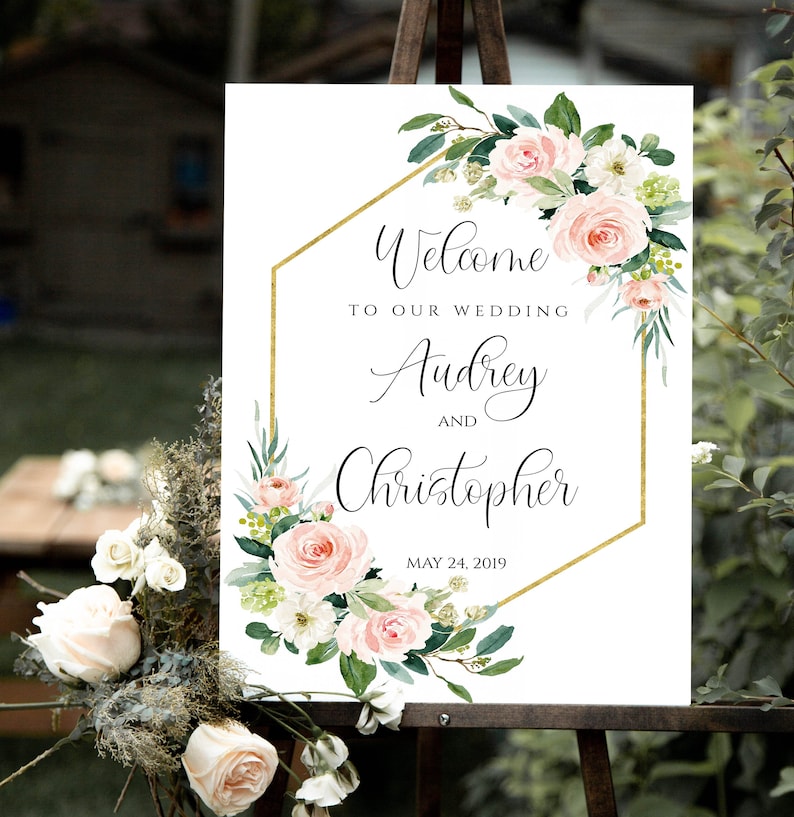 May include: A white wedding welcome sign with a gold hexagonal frame and pink and white floral design. The text reads "Welcome to our wedding Audrey and Christopher May 24, 2019".