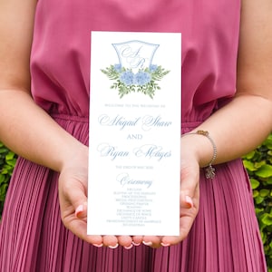 May include: A white wedding ceremony program featuring a blue crest with the initials AR, blue hydrangea floral illustrations, and text detailing the wedding of Abigail Shaw and Ryan Hayes on May 23rd, 2020, including the order of events.