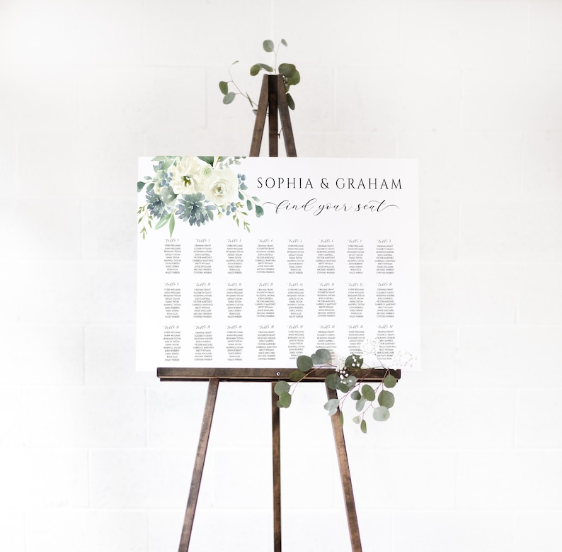 May include: A white wedding seating chart on a wooden easel, adorned with floral accents in shades of white, blue, and green. The text reads "Sophia & Graham find your seat."
