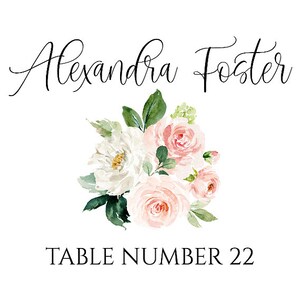 May include: A floral table number card with a white background. The card features a bouquet of pink and white flowers with green leaves. The text "Alexandra Foster" is written in cursive at the top of the card. The text "TABLE NUMBER 22" is written in black at the bottom of the card.