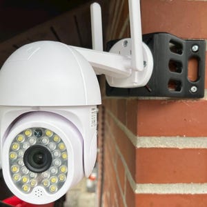 Puede incluir: Una cámara de seguridad blanca con una parte superior en forma de cúpula y un anillo de luces amarillas alrededor de la lente. La cámara está montada en un soporte negro fijado a una pared de ladrillo rojo. Dos antenas blancas se extienden desde la cámara.