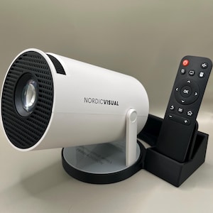 Op de afbeelding: Een witte cilindrische projector met een zwarte voorkant en de tekst "NORDICVISUAL". Een zwarte afstandsbediening en standaard staan rechts. De projector staat op een ronde basis.