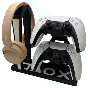 May include: A black and white gaming console stand holding two white and black game controllers and a tan headset. The stand has the letters and symbols "X O" on the base.