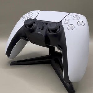 Peut inclure: Une manette PlayStation 5 blanche et noire sur un support noir. La manette présente un design épuré avec des boutons et des pavés directionnels. Le support maintient la manette en position verticale. L'arrière-plan est de couleur neutre.