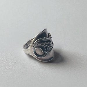 Puede incluir: Un anillo plateado con un diseño ornamental y arremolinado. El anillo presenta una sección curva y elevada con patrones detallados. El anillo se encuentra sobre un fondo liso y claro, resaltando sus intrincados detalles y artesanía.