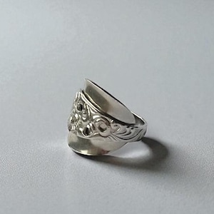 Puede incluir: Un anillo de plata hecho de una cuchara, con intrincados detalles florales y de volutas. El anillo tiene un diseño único, con el cuenco de la cuchara como elemento destacado. El anillo se muestra sobre un fondo blanco.