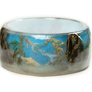 May include: A wide, resin ring with an underwater scene. The ring features a mermaid, seaweed, and a seashell against a blue background. The ring's outer edges have a brown, textured appearance.