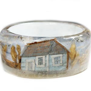 May include: A wide, clear resin ring with a painted landscape scene. The scene features a light blue house with a brown roof, set against a cloudy sky with birds. The ring's interior is white.