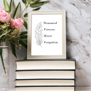 May include: A framed print with the words "Treasured Forever, Never Forgotten" and a feather illustration. The frame is light-colored and rests on a stack of books. Pink roses and greenery are in a vase to the left.