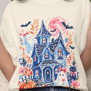 May include: A cream-colored short-sleeve t-shirt featuring a colorful graphic print of a blue haunted house, ghosts with pink bows, a black cat, a large orange pumpkin, bats, and a pink moon surrounded by autumn leaves and lit candles.