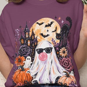 May include: A plum-colored t-shirt featuring a graphic print of a ghost wearing sunglasses and blowing a pink bubble, surrounded by a haunted house, a full moon, bats, a black cat, pumpkins, and flowers in shades of orange, purple, and black.