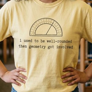 May include: A pale yellow short-sleeved t-shirt featuring a black line drawing of a protractor and the text "I used to be well-rounded then geometry got involved" printed in a typewriter-style font on the center of the chest.