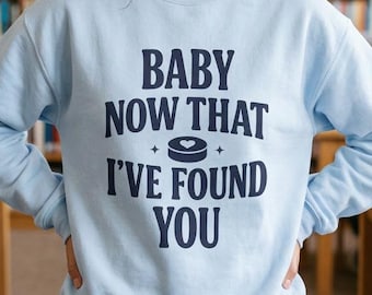 Könnte beinhalten: Ein hellblaues Langarm-Sweatshirt mit dunkelblauem Text, der BABY NOW THAT I'VE FOUND YOU lautet, und einer kleinen Eishockey-Puck-Grafik in der Mitte. Das Kleidungsstück hat einen Rundhalsausschnitt und gerippte Bündchen für einen schlichten Look.