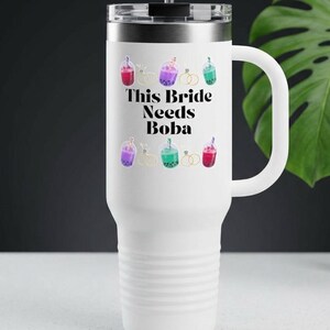 May include: White travel mug with a handle and a black lid. The mug features the text "This Bride Needs Boba" in black, surrounded by colorful boba tea illustrations and wedding rings. A black straw is inserted into the lid.