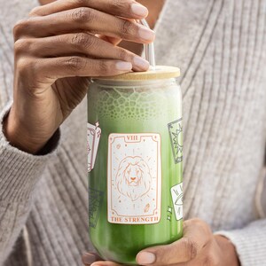 以下が含まれることがあります： 緑色の飲み物が入った、竹製の蓋とガラスのストローが付いた透明なガラスのタンブラー。タンブラーには、ライオンと「THE STRENGTH」の文字が入ったタロットカードのデザインが施されています。