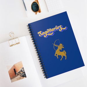Peut inclure: Un cahier à spirales bleu avec le mot "Sagittaire" en jaune. Une illustration dorée d'un centaure avec un arc et une flèche est sur la couverture. Le cahier est ouvert sur une surface blanche, avec un trombone doré.