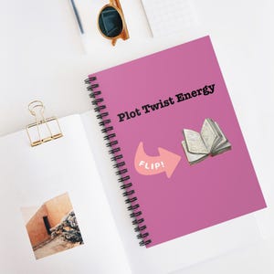 May include: A spiral-bound notebook with a magenta cover featuring the text "Plot Twist Energy." An illustration of an open book and a pink arrow with the word "FLIP!" are also on the cover. The notebook is on a white surface.