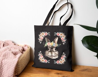 Peut inclure: Sac fourre-tout noir avec un motif de lapin portant des lunettes et une couronne de fleurs. Le lapin est entouré de compositions florales. Le sac a des anses noires et est fait d'un tissu durable, idéal pour les courses ou les livres.