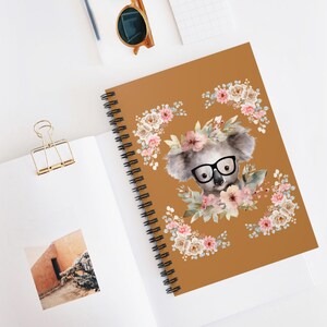 Peut inclure: Un cahier à spirales avec une couverture marron représentant une illustration à l'aquarelle d'un koala portant des lunettes et une couronne de fleurs. Le koala est entouré de fleurs roses et crème. Le cahier est ouvert sur une surface blanche.