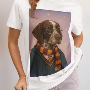 May include: White t-shirt featuring a dog portrait. The dog, wearing glasses and a scarf, is depicted in a wizard-like style. The background has a starry effect. The shirt is a casual, everyday wear item.