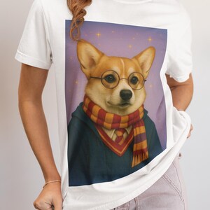 May include: White t-shirt featuring a corgi wearing round glasses, a striped scarf, and a blue robe. The corgi is set against a purple starry background. The shirt is a casual, everyday wear item.