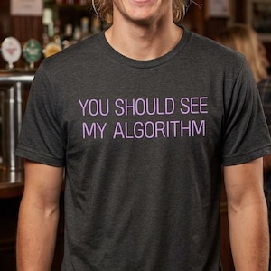 May include: Dark gray t-shirt with the text "YOU SHOULD SEE MY ALGORITHM" in purple. The shirt is worn by a person smiling in a bar setting. Other people are in the background.