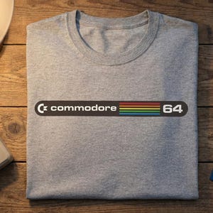 Może przedstawiać: Szara koszulka z napisem "Commodore 64" w czarnym prostokącie z tęczowym wzorem w paski. Koszulka ma okrągły dekolt. Projekt jest wyśrodkowany na przodzie koszulki. Koszulka wykonana jest z miękkiego materiału.