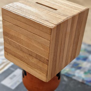 Könnte beinhalten: Eine würfelförmige Holzspardose mit einem Schlitz zum Einwerfen von Geld. Die Spardose besteht aus horizontalen und diagonalen Holzlatten, die ihr ein strukturiertes Aussehen verleihen. Sie steht auf einem terrakottafarbenen Sockel.