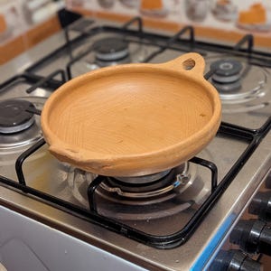 以下が含まれることがあります： ガスコンロのバーナーの上に置かれた、取っ手付きの丸い素焼きの調理皿。皿は薄茶色で、手作りのようです。コンロはステンレス製です。