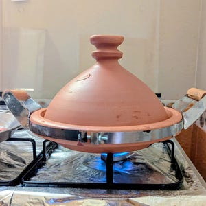 Pode incluir: Uma panela de tagine de terracota com tampa cónica, colocada num fogão a gás. A panela tem uma faixa e pegas de metal. O fogão está ligado, com uma chama azul visível por baixo.