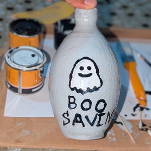 Op de afbeelding: Een wit geschilderde vaas met een spookillustratie en de woorden "BOO SAVIN" in het zwart. De vaas wordt door een hand vastgehouden. Verfblikken en een penseel staan op de achtergrond.